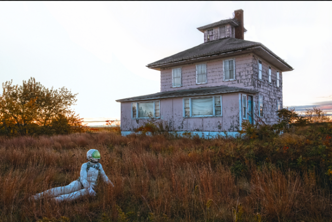 An abandoned weathered two-story house sits in an overgrown field at sunset, with an astronaut figure lying in the tall grass in the foreground.