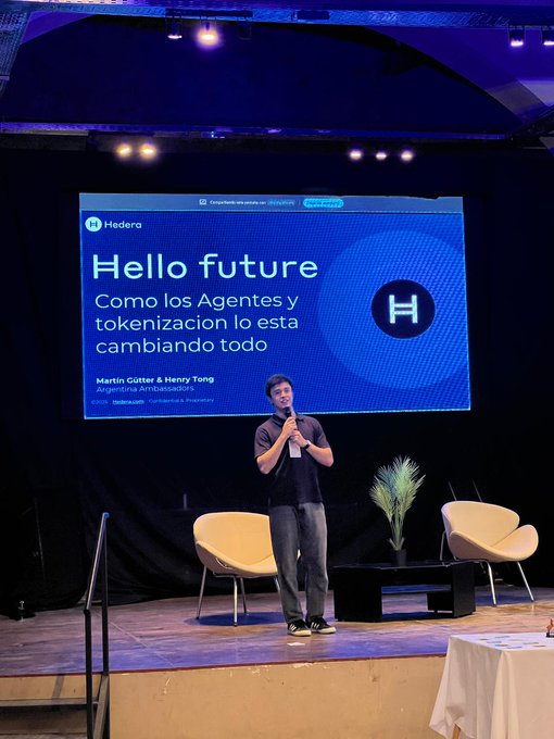 In the image, a man stands confidently at a podium with a large screen behind him displaying "Hello Future". He appears to be giving a presentation or speech related to the community growth of Hedera. The room has a stage setup with chairs arranged around the area where he is speaking. There are also potted plants placed on either side of the stage, adding some greenery and visual interest to the scene.