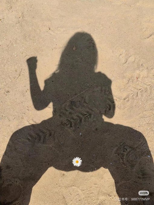 A shadow of a person sitting in sand with one arm raised, with a small white daisy emoji placed at the crotch area.