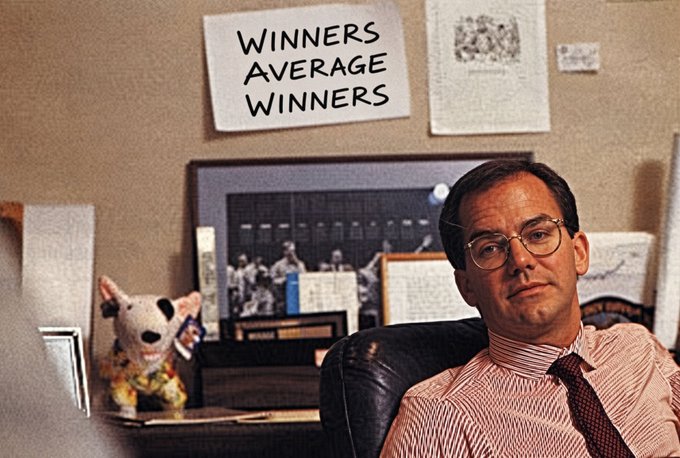 A man in glasses and a striped shirt with a tie sits at a desk in an office; behind him is a sign reading 'WINNERS AVERAGE WINNERS' and a Spuds Mackenzie figurine.