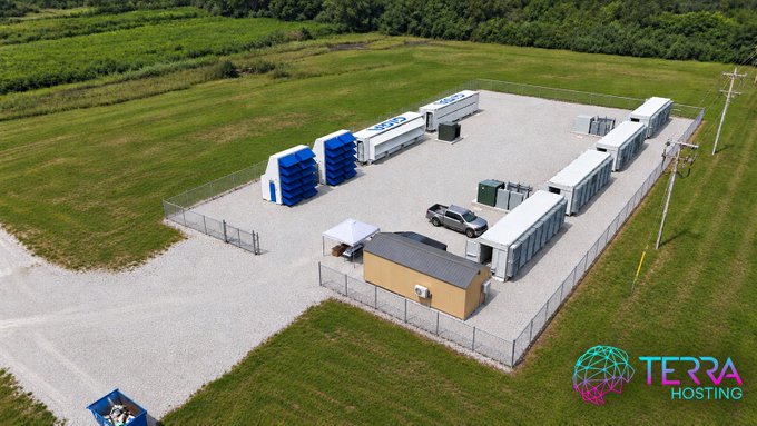An aerial drone photograph of a fenced industrial data center compound featuring shipping container-style server modules, blue cooling units, and a small building, branded with 'TERRA HOSTING' logo in the corner.
