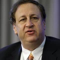 A headshot of a middle-aged man with brown hair wearing a dark suit, white shirt, and orange tie, appearing to speak in a formal setting.