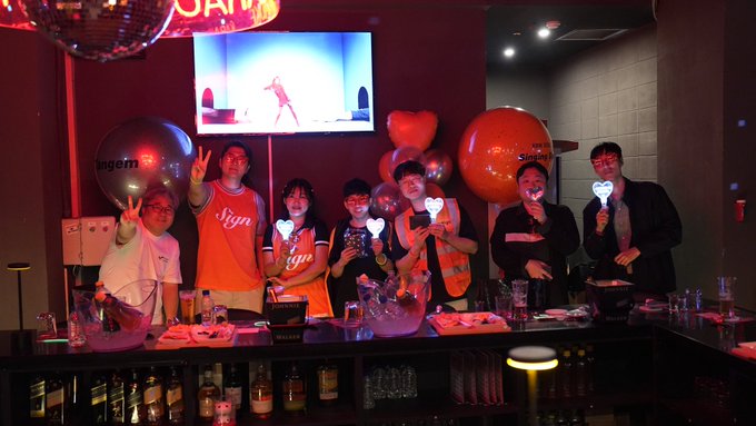 A group of seven people posing behind a bar with glowing heart-shaped signs, balloons branded with "Singtel," and a TV screen in the background at what appears to be a corporate event.