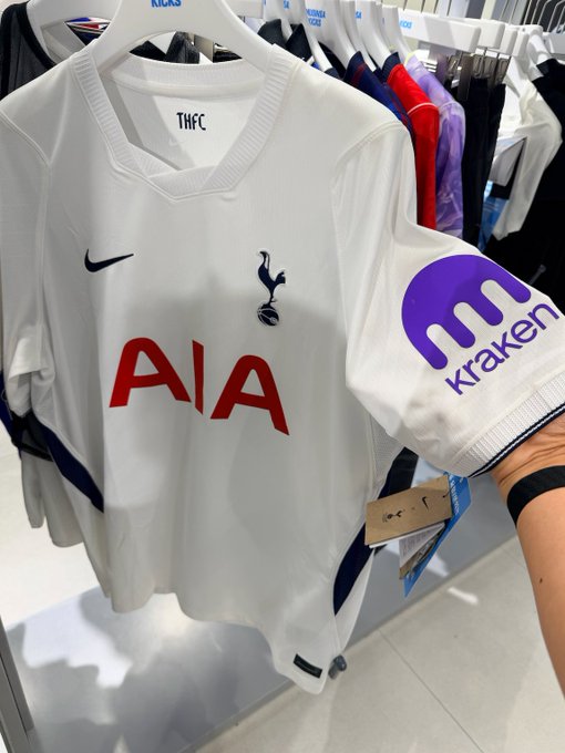 A Tottenham Hotspur FC (THFC) white Nike football jersey featuring the AIA sponsor logo and a Kraken cryptocurrency patch on the sleeve hangs on a hanger in a store, with Nike and other jerseys visible in the background.
