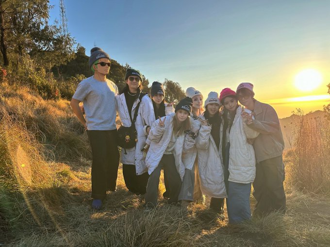 Seven friends gather on a grassy hilltop at sunrise, bundled in winter coats and beanies, posing together in a group photo. The caption celebrates Mid-Autumn Festival, matching the warm, communal atmosphere captured as the sun rises behind them.