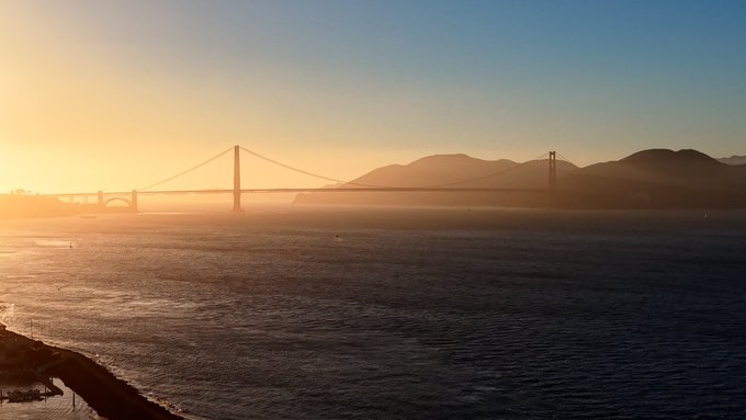 A golden sunset bathes a suspension bridge spanning a calm bay, with silhouetted mountains visible in the background.