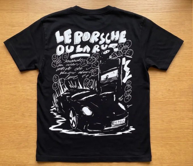 A black t-shirt laid flat on a wooden surface, featuring white hand-drawn artwork with the text 'LE PORSCHE OU LA RUE' and an illustration of a sports car.