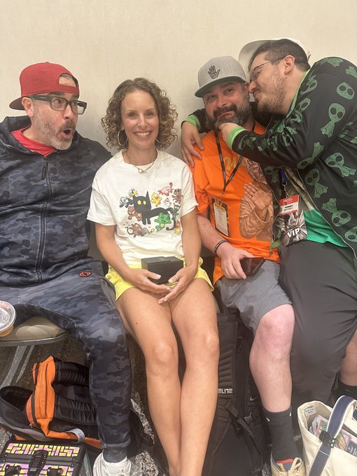 Four people pose together on a couch, appearing playful and happy; from left, a man in a camo hoodie with a surprised expression, a woman holding a black object, a man in an orange shirt, and a man in a green patterned hoodie giving a side glance.