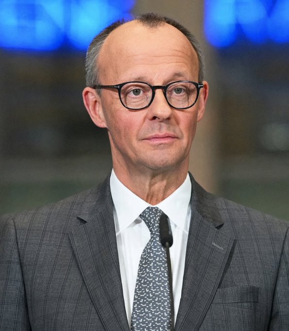 A portrait of a man with short gray hair and black-rimmed glasses, wearing a gray suit with a white shirt and patterned tie, photographed against a blurred blue background.