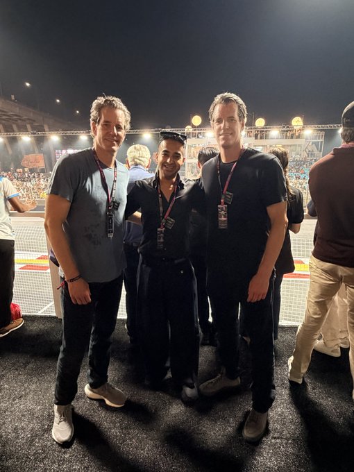 Three men pose together at what appears to be a racing event at night, with stadium lights illuminating a crowded grandstand in the background. The two men on either side wear lanyards and casual attire, while the man in the middle sports a black shirt, fitting the post's caption highlighting notable Bitcoin figures Tyler and Cameron, reportedly pictured alongside over 100,000 BTC combined.