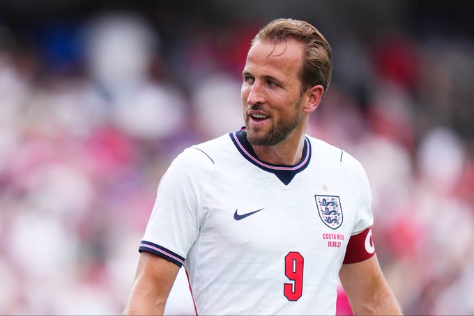 The image features a soccer player wearing an England jersey, standing on a field with his arms crossed. The background of the photo shows a blurred crowd watching the game from behind him. There are also several other people present in the scene, but they are not as prominent as the main subject. The post is titled "$250 to four if Harry Kane has a shot on target for England tonight - just repost & follow @DuelbitsSports". This suggests that the image might be related to a sports event or match between England and another team, possibly in the context of a soccer game.