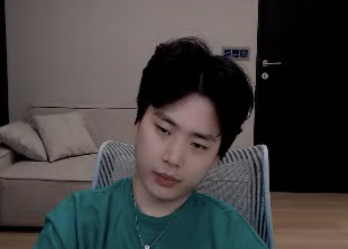 A young man with dark hair wearing a teal shirt sits in a striped chair in a living room, looking sideways with a contemplative or skeptical expression that mirrors the cautious sentiment of the post advising against aggressive BTC trading until the micro point of control at 64.2k is reclaimed.