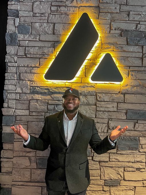 A smiling man in a black blazer, white shirt, and a cap standing in front of a stone wall with an illuminated Avalanche (AVAX) logo.
