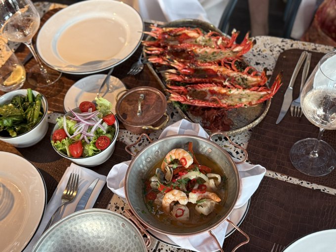 An overhead view of a dining table featuring a seafood spread including a large platter of prawns, a pot of seafood soup, a fresh salad, and greens, accompanied by wine glasses.