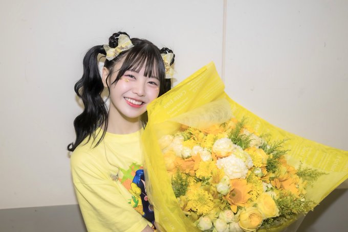 A smiling young woman with pigtails and yellow hair ribbons holds a large bouquet of yellow and white flowers wrapped in yellow paper.