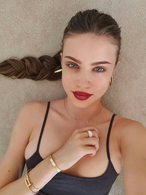A portrait of a young woman with braided brown hair, wearing a black top, red lipstick, and gold jewelry, lying down and looking directly at the camera.