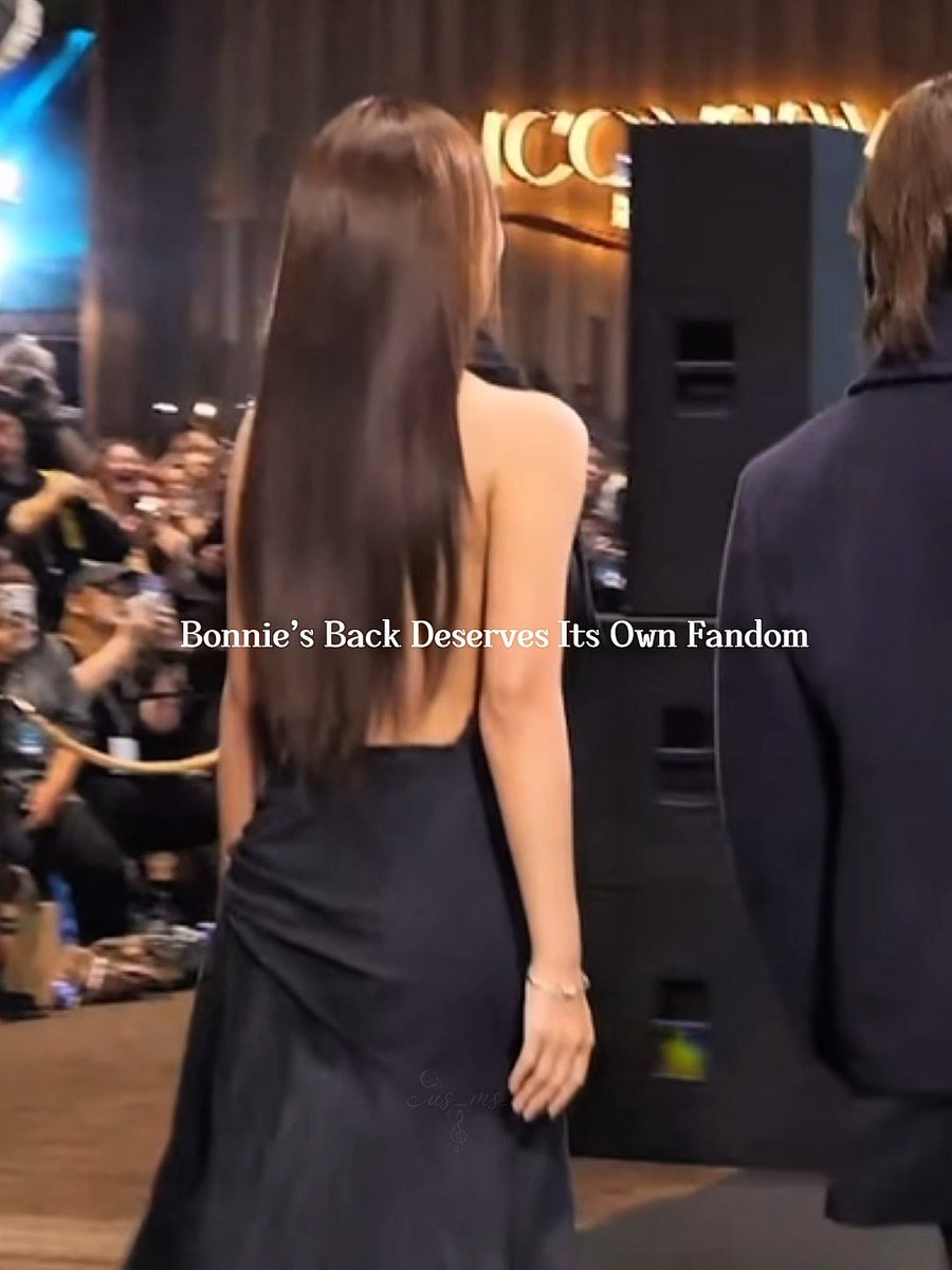 A woman with long brown hair walks on stage wearing an elegant backless black gown at the M Choice Mint Awards 2026, while fans photograph her from the sidelines; overlaid text reads "Bonnie's Back Deserves Its Own Fandom," highlighting fans' admiration for Emi Bonnie's striking red carpet look.