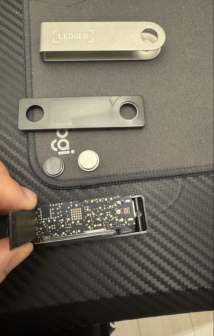A hand holds a Ledger hardware wallet device with its protective metal case and a small envelope resting on a carbon fiber-textured surface.