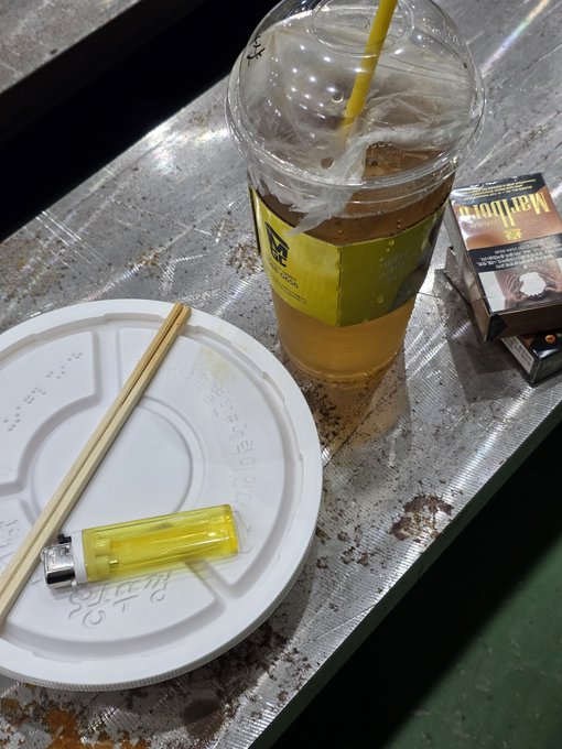 A meal setting on a metallic tray featuring a partially consumed iced drink in a clear plastic cup with a yellow straw, an empty white plate with wooden chopsticks and a yellow lighter, alongside a Marlboro cigarette pack.