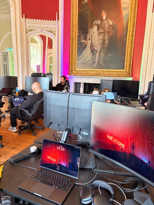 An ornate room with red walls and a large classical portrait contains a workspace with multiple monitors and laptops displaying 'WELCOME' screens.
