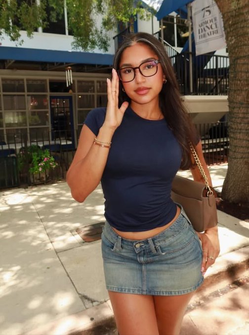 A young woman in glasses, a navy fitted top, and denim mini skirt poses on a sidewalk in front of a building with a banner reading "15 Armour Dance Theatre," giving a casual street-style wave to greet followers with the "gm" (good morning) energy. The posed, stylish shot fits the relaxed, confident tone of a weekly "gm" check-in seeking crypto alpha.