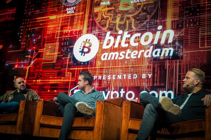 Three men sit on a stage beneath a large screen reading “bitcoin amsterdam” and “PRESENTED BY vanta.com.” The image accompanies a giveaway announcement for Michael van de Poppe’s appearance and five Bitcoin Amsterdam conference passes.