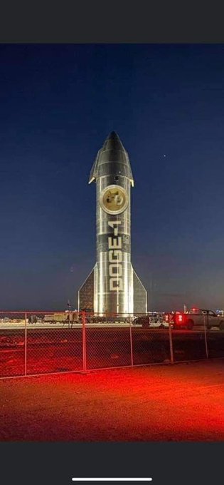 Nighttime photograph of a Dodge rocket-shaped display or monument illuminated against a dark blue sky, with a red emergency vehicle parked nearby and a chain-link fence in the foreground.