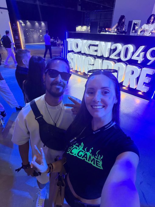 Two people pose for a photo at an event with a neon sign reading 'TOKEN 2049 SINGAPORE' in the background, illuminated by blue lighting.