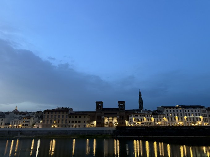 A serene twilight view of buildings along a river, with warm lights reflecting on the water under a deep blue cloudy sky.