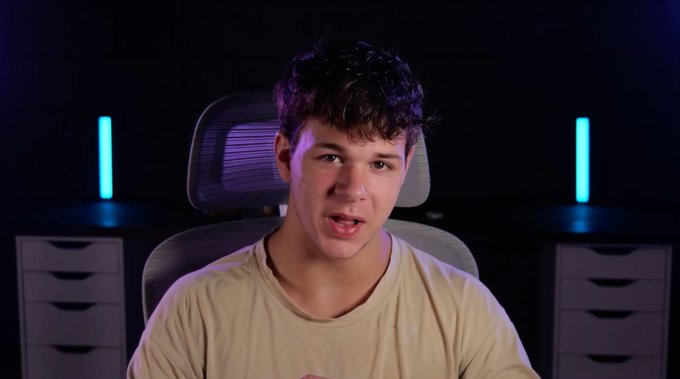 A young man in a beige t-shirt sits in an office chair against a dark backdrop featuring two glowing blue light bars, appearing to speak directly to the camera in what looks like a content creator's recording setup for an upcoming video announcement about the cryptocurrency $CATE.