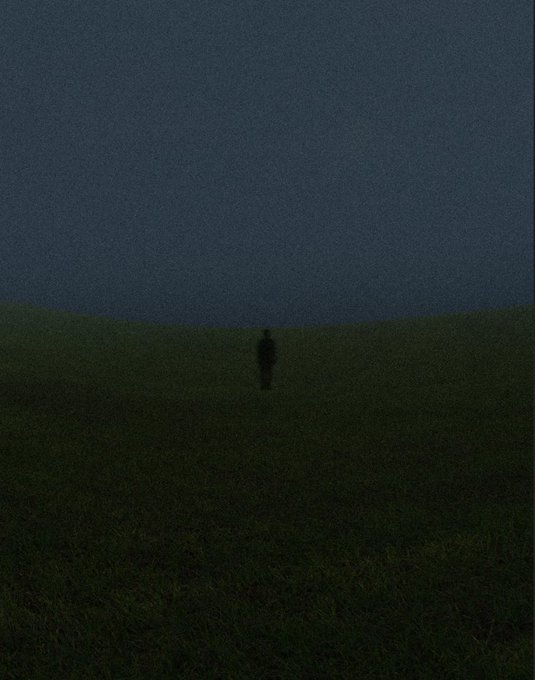 A solitary silhouetted figure stands in the middle of a vast grassy field under a dark, moody sky.