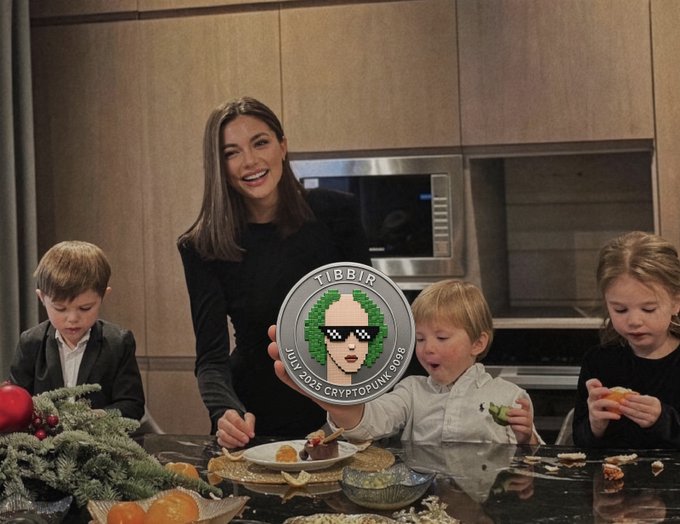 A smiling mother and three children gather around a kitchen counter displaying a large $TIBBIR (CryptoPunk #9588) coin, playfully referencing the joke about the family becoming wealthy after dad bought and held the $TIBBIR token to "tibbirillions." The scene captures a humorous "rich family" moment centered around the cryptocurrency meme coin.