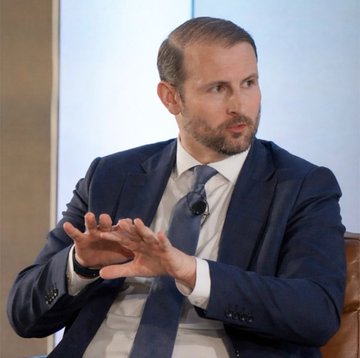 A man in a dark suit and blue tie gestures with both hands while speaking during what appears to be an interview or discussion. The image illustrates the accompanying news post about Patrick Witt, the White House's Advisor of Digital Assets, expressing concerns that Democrats prevented a vote on the Clarity Act before the August recess.