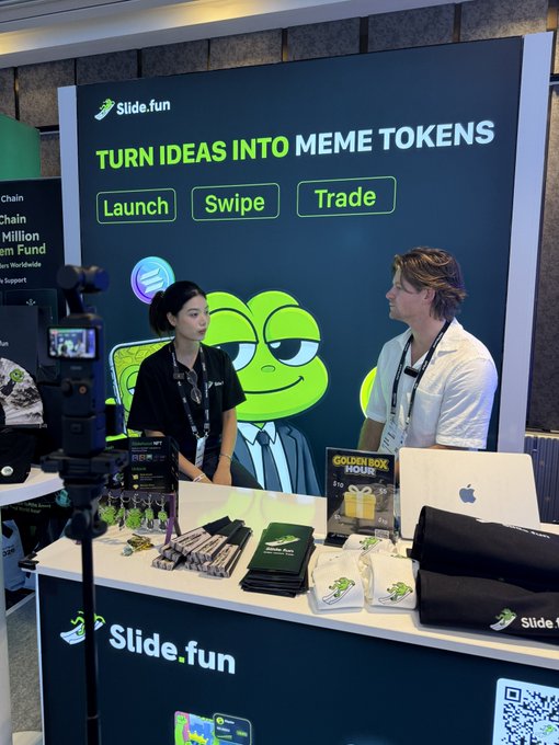 Two people stand at a 'Slide.fun' booth with a banner reading 'TURN IDEAS INTO MEME TOKENS' and buttons for 'Launch', 'Swipe', and 'Trade', with merchandise including 'Golden Box Mouse' items on the counter.