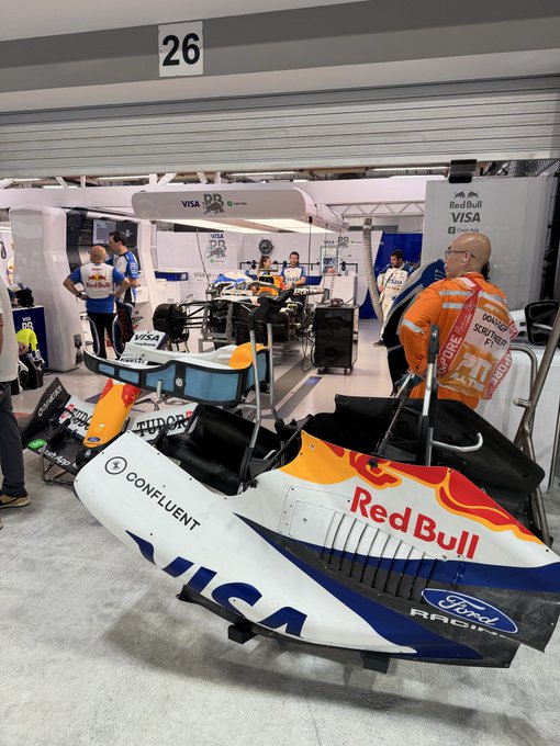 A Red Bull Racing Formula 1 car in the garage area at a race circuit with crew members in Oracle Red Bull Racing uniforms.