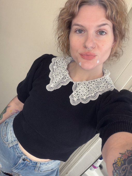 A curly-haired woman with a nose ring takes a selfie wearing a black knit top featuring a striking white lace Peter Pan collar, with visible tattoos on her arm. The image playfully accompanies her post about being accused of witchcraft, with the ornate vintage-style lace collar reinforcing the historical 1692 Puritan-era aesthetic referenced in her tongue-in-cheek crypto joke.