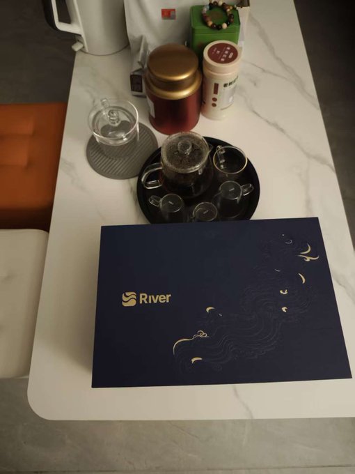 A tea set with a glass teapot on a tray sits on a marble table next to a navy blue 'River' branded folder with gold wave designs, alongside cookies and a kettle.