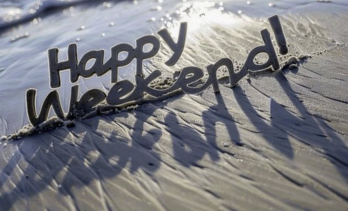 The phrase "Happy Weekend!" is written into wet sand at the beach, with sunlight reflecting off the water and casting shadows of the letters.