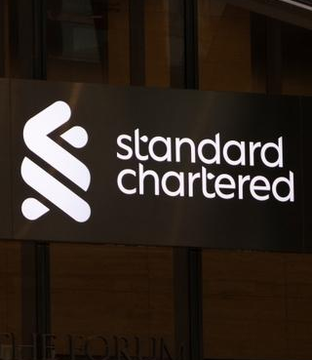 A Standard Chartered bank sign is displayed on a building entrance, featuring the company's logo and name. The image relates to the post as Standard Chartered's research head, Geoff Kendrick, has predicted that Bitcoin's year-end target of $100K is "too low," suggesting an accelerated BTC rally.