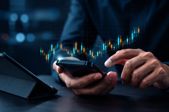 A person holds a smartphone displaying a candlestick financial chart with orange and teal bars hovering above the screen, suggesting stock trading or market analysis.
