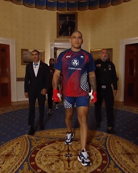 The image shows a man walking into an ornate room with a large circular rug, while several people are standing around him. The man appears to be wearing a UFC uniform and carrying boxing gloves. In the background, there are two men dressed in suits, one of whom is holding a tie. A third person can also be seen in the scene. The room has a white door on the left side and a painting hanging above it. The image seems to be related to an event or gathering where the man in the UFC uniform is walking into the room as a first responder, possibly attending to an emergency situation or providing assistance.