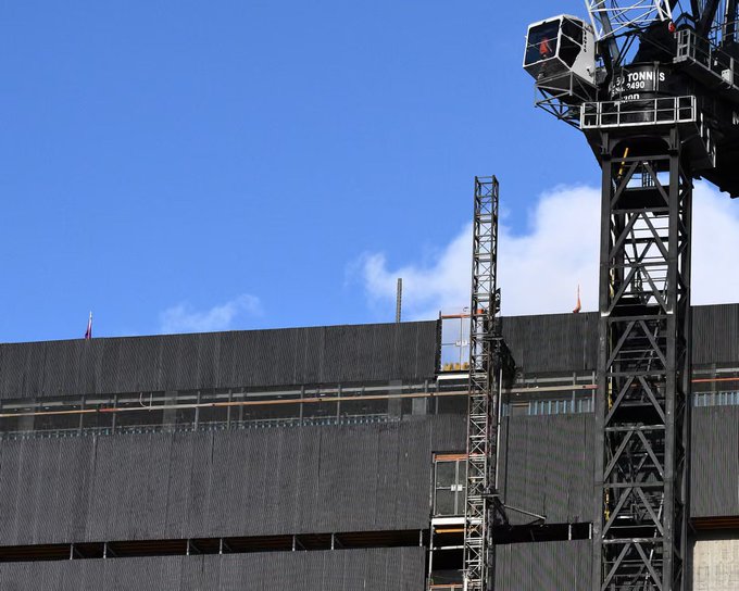 A "75 TONNES" tower crane rises above a large industrial facility under construction against a blue sky, symbolizing the massive infrastructure buildup referenced in the post about a $7 trillion AI investment and the urgent need for accessible compute—exactly the gap that decentralized infrastructure aims to solve.