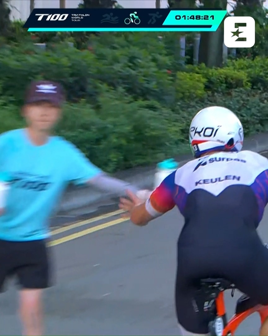 A cyclist labeled "KEULEN" on their suit receives a water bottle from a runner in a teal T100 shirt, with the T100 Triathlon World Tour race clock showing 01:48:21, capturing the teamwork between biker and runner.