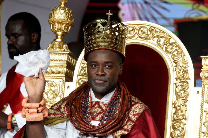 A man dressed as a Nigerian king or chief sits on an ornate gold throne wearing a gold crown with a cross, traditional coral beads, and rich red royal attire, while holding a white handkerchief.