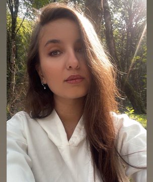 Portrait selfie of a young woman with long brown hair wearing a white shirt, standing outdoors with trees in the background.