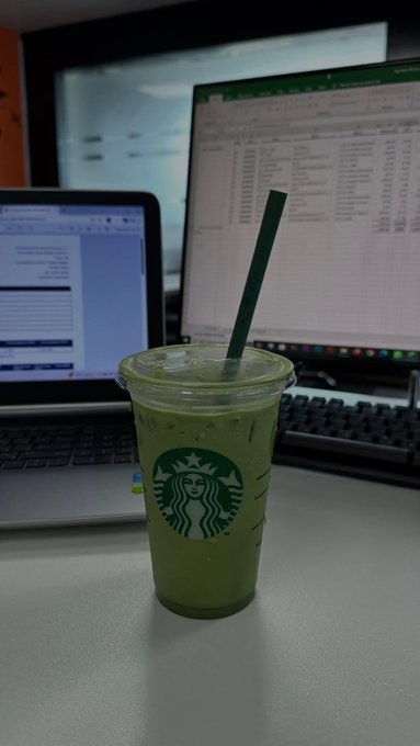 A Starbucks iced green tea matcha latte in a clear plastic cup with a green straw sits on a desk in front of a laptop and an external monitor displaying spreadsheets.