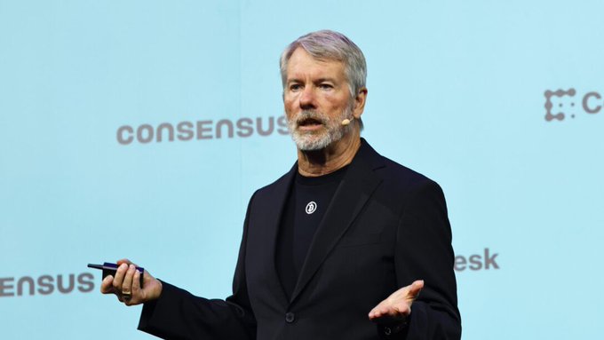 A man in a black blazer speaking at the Consensus conference, wearing a lapel pin and holding a clicker, with the 'consensus' logo visible in the background.