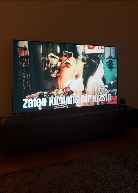 A television mounted on a dark console displays a scene from a show or movie featuring a person with sunglasses, with Turkish subtitles reading 'Zaten Kurtulmuş Bir Kizsin'.