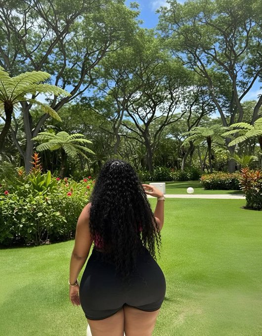 Photo from behind of a woman with long curly hair taking a picture in a lush tropical garden with tall trees and green lawn.