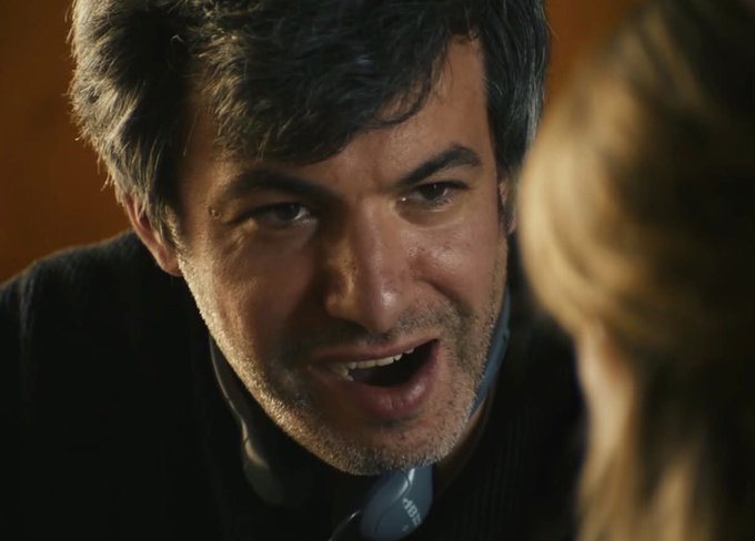 A close-up of a man with dark hair and stubble, wearing a dark sweater over a light blue shirt, speaking with an intense expression while looking at someone off-camera.
